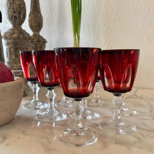 May include: Set of eight red wine glasses with clear stems and bases. The glasses have a cut glass design and are arranged on a white marble surface.