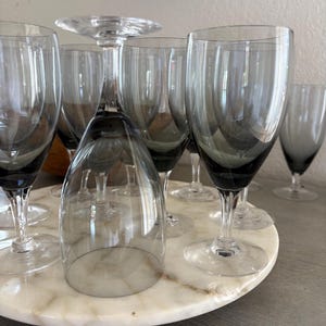 May include: A collection of gray-tinted wine glasses with clear stems and bases, arranged on a round, marble-patterned serving tray. One glass is inverted. The glasses are of varying heights and shapes, suggesting a set for different wines.