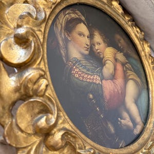 May include: Oval-shaped, antique-style framed artwork. The frame is ornate, gold-colored, and features a classical painting of a woman and child. The painting has a dark background and the figures are dressed in period clothing.