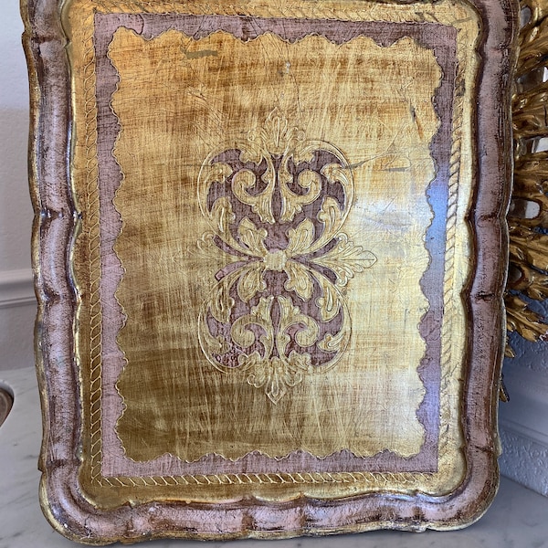 Puede incluir: Una bandeja decorativa con acabado dorado y rosa. La bandeja tiene un diseño ornamentado en relieve en el centro y un borde festoneado.