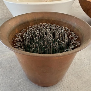 May include: A copper-colored flower frog with a metal grid, filled with green material, sits on a gray surface. A white ceramic mortar and wooden pestle are in the background, along with a wooden bowl.