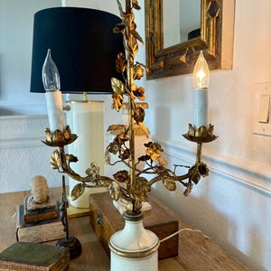 Może przedstawiać: Złota metalowa lampa kandelabrowa z dwoma oprawkami i białymi osłonami świec, ozdobiona misternymi metalowymi motywami kwiat&oacute;w i liści. Lampa stoi na okrągłej, złamanej bieli podstawie ze złotym wykończeniem i posiada uchwyt w kształcie pierścienia.