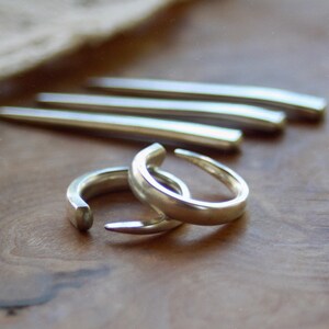 May include: Two silver-colored open rings with a smooth, polished finish and tapered ends, positioned in the foreground. Behind the rings, three elongated, slightly curved silver-colored metal rods are arranged horizontally in a row.