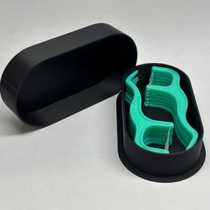 May include: A black, oval-shaped container with a lid, holding several green dental floss picks. The container is open, revealing the floss picks neatly arranged inside. The background is a plain white surface.