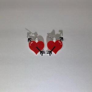 May include: A pair of heart-shaped earrings. Each red heart has a black arrow piercing through it. The earrings have silver-colored hooks. The earrings are a romantic accessory.
