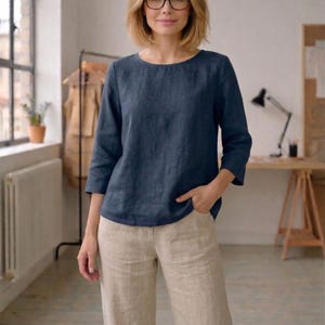 May include: A woman wearing a navy blue linen top with three-quarter sleeves and beige linen pants. The top has a round neckline. The woman is wearing glasses. The background includes a window, a desk, and a clothing rack.
