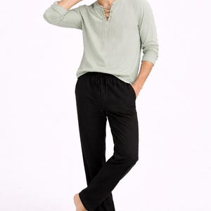 Puede incluir: Una camisa verde claro de manga larga con cuello con cordones y pantalones negros con cordón. La camisa tiene un corte relajado y los pantalones son de pierna recta. El atuendo es informal y cómodo.