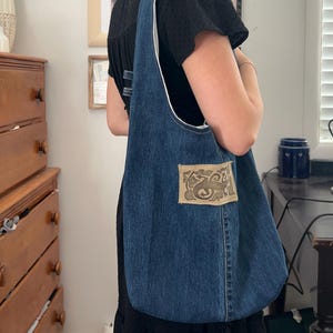 Op de afbeelding: Een denim tote bag met een witte binnenvoering en een decoratieve patch. De tas is van een medium blauwe kleur en heeft een lange band. De patch heeft een sierlijk ontwerp in tinten beige en bruin.