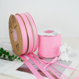Puede incluir: Carretes y rollos de cinta rosa. La imagen muestra varios carretes de cinta rosa, con algunos hilos de cinta desatados. La cinta es de un color rosa pastel suave. Los carretes son de cartón y plástico.