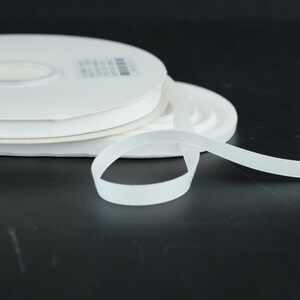 May include: White elastic ribbon spools. The ribbon is unwound from the spool, showing the flat, soft texture. The spools are stacked on top of each other. The background is a dark, solid color.