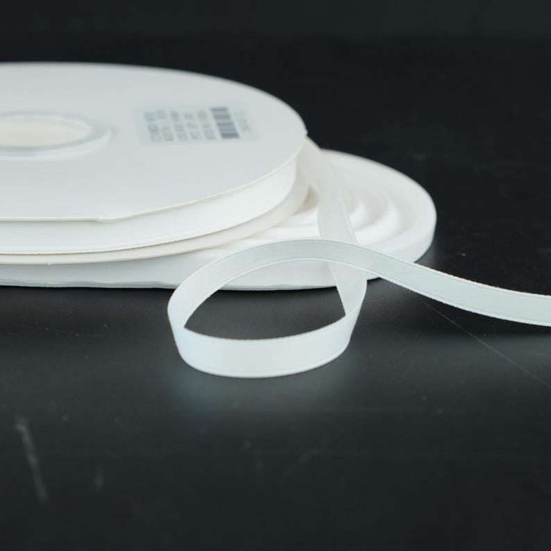 May include: White elastic ribbon spools. The ribbon is unwound from the spool, showing the flat, soft texture. The spools are stacked on top of each other. The background is a dark, solid color.
