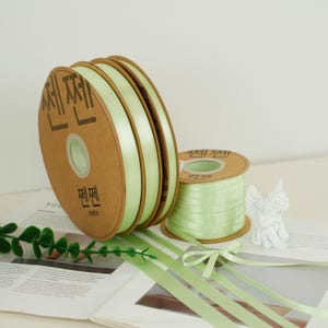 May include: Several spools of light green satin ribbon are displayed, with a small spool and a few larger ones. The spools are made of cardboard and have Korean text printed on them. A small white angel figurine is also present.