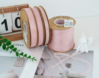 May include: Several spools of pale pink ribbon are displayed, with a few spools stacked together and one standing alone. The ribbon is a sheer, delicate material. A small white cherub statue and a calendar are also visible in the background.