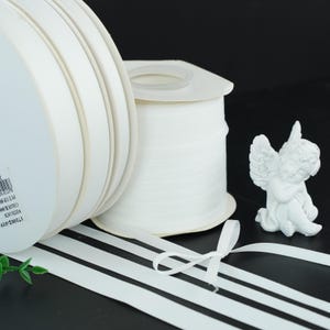 May include: White satin ribbon rolls and a small white angel figurine. The ribbon is unwound, with a small bow tied in the foreground. The rolls of ribbon are stacked, and the angel is resting on a black surface.