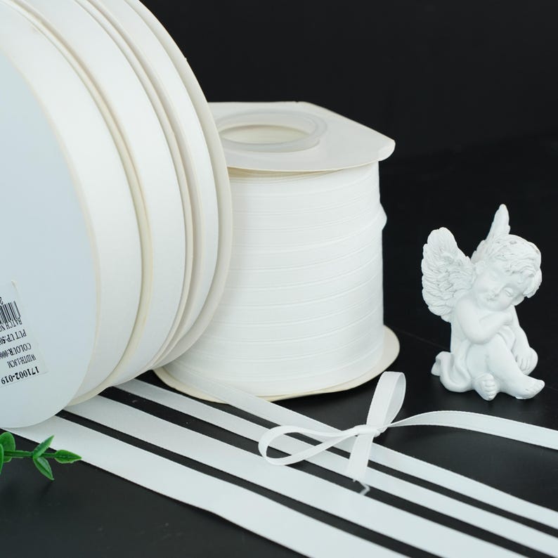 May include: White satin ribbon rolls and a small white angel figurine. The ribbon is unwound, with a small bow tied in the foreground. The rolls of ribbon are stacked, and the angel is resting on a black surface.