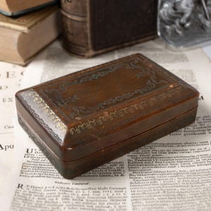 May include: A rectangular brown wooden box with a hinged lid, featuring intricate gold-colored filigree patterns along the edges and a decorative border design on the top surface, showcasing a smooth, polished finish.