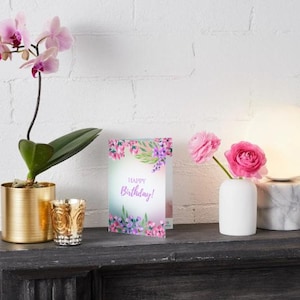 May include: A still life arrangement featuring a gold planter with a pink orchid, a decorative candle holder, and a greeting card that says "Happy Birthday!". A white vase holds pink roses, and a round lamp sits on a marble base.