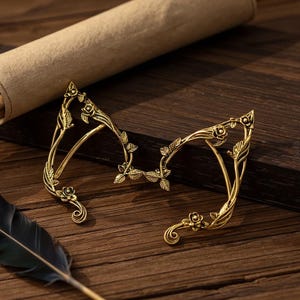 May include: A pair of gold-colored elf ear cuffs with floral and leaf detailing. The ear cuffs are made of metal and are designed to be worn on the ears. The cuffs are placed on a wooden surface with a feather and a rolled-up scroll in the background.
