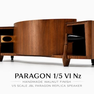 Puede incluir: Una réplica de altavoz JBL Paragon a escala 1/5 con acabado en nogal. La unidad presenta un frente de madera curvado, dos compartimentos laterales con interiores negros, un acento ovalado dorado a la izquierda y un cono negro a la derecha. Texto: PARAGON 1/5 VI Nz HANDMADE WALNUT FINISH 1/5 SCALE JBL PARAGON REPLICA SPEAKER.