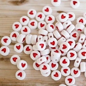 May include: A large pile of white heart-shaped beads with red centers. The beads are arranged in a random pattern and are all the same size.