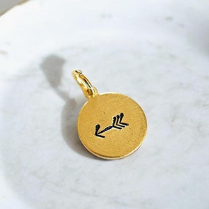 May include: Gold-colored charm with a black arrow pointing to the left. The charm is round and has a loop for attaching to a necklace or bracelet.