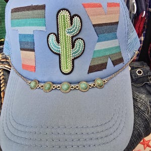 Op de afbeelding: Lichtblauwe truckerpet met de letters "TX" in een gestreept ontwerp en een cactuspatch. Een zilveren ketting met turkooizen stenen is over de rand gedrapeerd. De pet heeft een mesh achterkant.