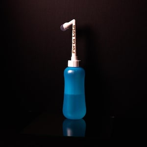 以下が含まれることがあります： 白いノズルと「Porta Lota」の文字がプリントされた青いプラスチックボトル。ボトルには透明な液体が入っており、曲線的なデザインです。ノズルには小さな透明な先端が付いています。