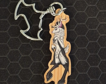 May include: A wooden keychain featuring a stylized illustration of a white deer with black markings and antlers. The deer is depicted in a standing pose. The keychain includes a silver-colored metal clasp shaped like a bat silhouette.