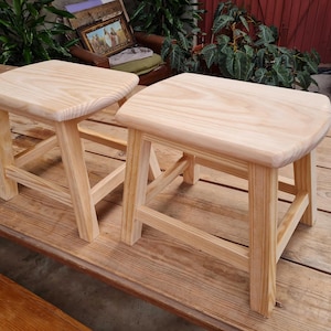 Puede incluir: Dos taburetes pequeños de madera clara con un acabado de veta natural. Cada taburete cuenta con un asiento rectangular de esquinas redondeadas y cuatro patas robustas conectadas por barras de soporte horizontales en todos sus lados.
