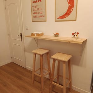 Op de afbeelding: Een aan de muur gemonteerde bar-tafel van licht hout met twee bijpassende hoge houten krukken. Boven de tafel hangen twee ingelijste prints: een met vijf rode sterren en de tekst "CHEF WAS CUTE, WOULD EAT HERE AGAIN." en een andere met een rode chilipeper.