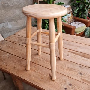 Puede incluir: Un taburete de madera clara con un asiento redondo y cuatro patas. El taburete está hecho de madera sin terminar, mostrando la veta natural. Las patas están conectadas por soportes horizontales. El taburete está sobre una mesa de madera.