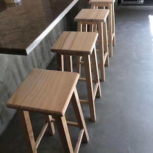 Könnte beinhalten: Eine Reihe von vier hellen Holzbarhockern mit quadratischen Sitzen und vier geraden Beinen. Jeder Hocker weist eine natürliche Holzmaserung auf und hat ein einfaches, funktionales Design mit horizontalen Stützstreben zwischen den Beinen.
