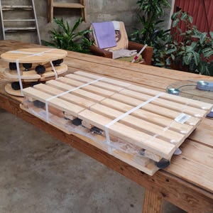 Op de afbeelding: Een rechthoekige houten plantenroller met latten en zwarte zwenkwielen, vastgezet met witte plastic banden. Daarnaast staat een ronde houten plantenroller, ook voorzien van zwarte zwenkwielen en vastgezet met witte plastic banden.