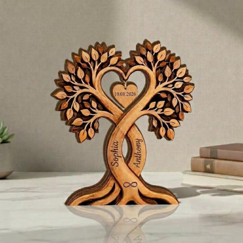 Op de afbeelding: Een houten boomsculptuur met twee verstrengelde stammen die een hartvorm vormen. Een klein hartvormig hangertje hangt in het midden met de tekst 10.08.2026. De namen Sophia en Anthony zijn in de stammen gegraveerd, samen met een oneindigheidssymbool.