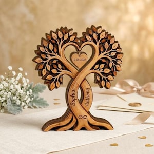 Op de afbeelding: Een houten boomsculptuur met twee verstrengelde stammen die een hartvorm vormen. De stammen zijn gegraveerd met de namen Sophia en Anthony en een oneindigheidssymbool. Een hartvormige hanger in het midden toont de datum 10.08.2026.