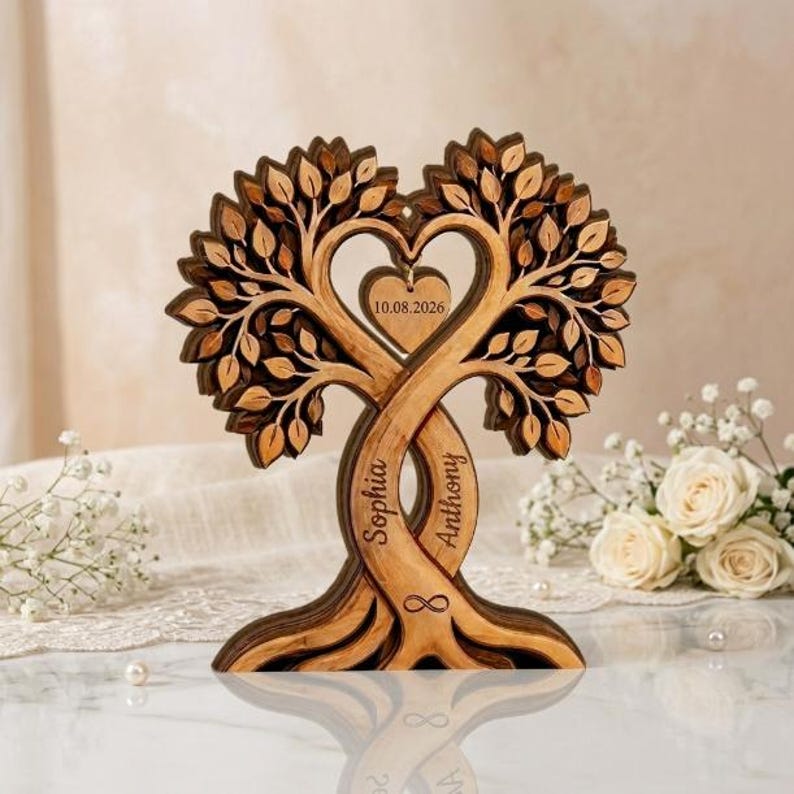 Op de afbeelding: Een gelaagd houten sculptuur met twee verstrengelde stammen die een hartvorm vormen. De stammen zijn gegraveerd met de namen Sophia en Anthony, en een centraal hartvormig hangertje toont de datum 10.08.2026 met een oneindigheidssymbool.