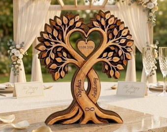 Op de afbeelding: Een houten boomsculptuur met twee verstrengelde stammen die een hartvorm vormen. De stammen zijn gegraveerd met de namen Sophia en Anthony en een oneindigheidssymbool. Een hartvormige hanger in het midden toont de datum 10.08.2026.