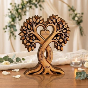 Op de afbeelding: Een houten boomsculptuur met twee verstrengelde stammen gegraveerd met de namen Sophia en Anthony, een centrale hartvormige uitsparing en een hangend hartornament met de datum 10.08.2026 en een oneindigheidssymbool aan de voet.