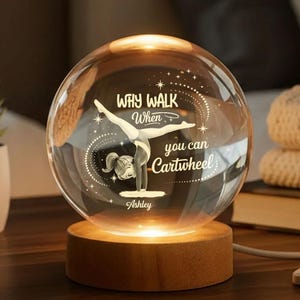 May include: A clear glass sphere with a gymnast illustration and the text "Why walk when you can cartwheel." The sphere sits on a wooden base that emits a warm glow. The gymnast is wearing a leotard and is doing a handstand.