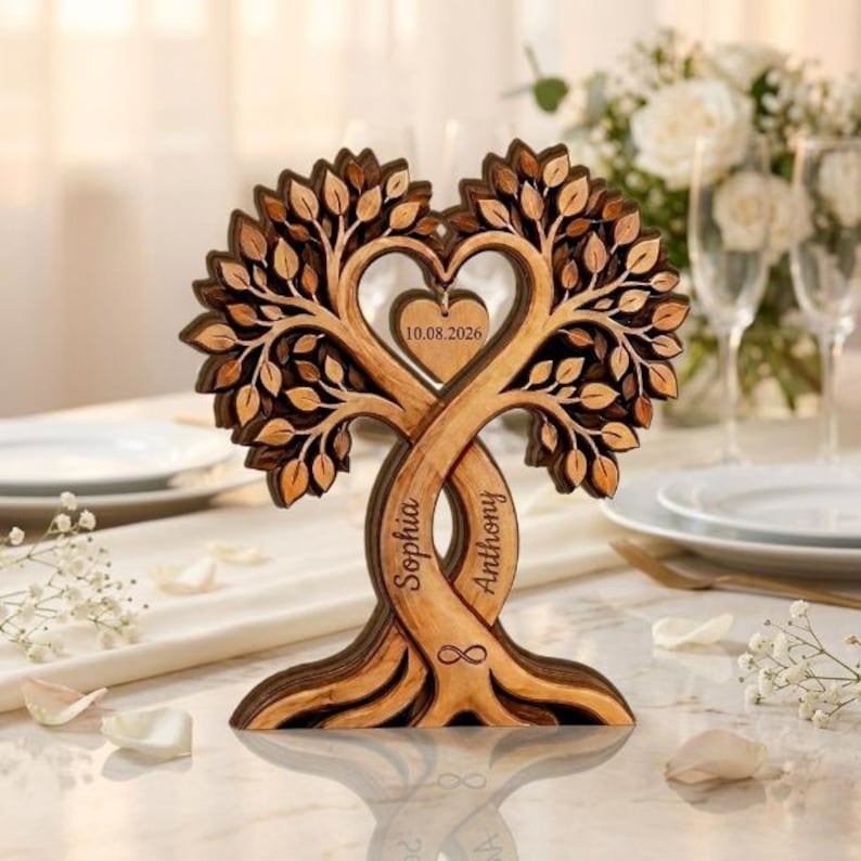 Op de afbeelding: Een gelaagd houten boomsculptuur met twee verstrengelde stammen die een hartvorm vormen. De stammen zijn gegraveerd met de namen Sophia en Anthony, en in het midden hangt een klein hartvormig hangertje met de datum 10.08.2026.