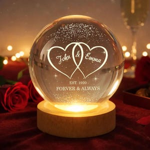 May include: A clear glass globe with two intertwined heart shapes, each bearing a name, and the words "Forever & Always." The globe sits on a wooden base, illuminated from within. The text "Est. 1920" is also visible.
