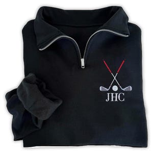 May include: Black golf pullover with a silver zipper and embroidered golf club design. The design includes crossed golf clubs, a golf ball, and the initials "JHC" in white. The sleeves are folded up.