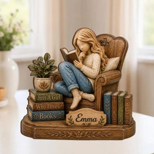 May include: A resin figurine of a person with long hair sitting in a wooden chair reading a book. The base features a stack of three books labeled Just A Girl Who Loves Books and a nameplate reading Emma, with a potted plant and additional books.
