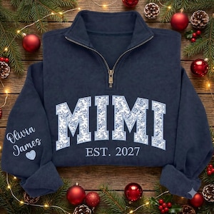 May include: A navy blue quarter-zip sweatshirt featuring the text MIMI in a white and blue floral pattern across the chest, with EST. 2027 printed below. The left sleeve displays the names Olivia James above a small light blue heart icon.