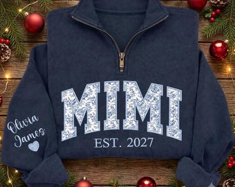 May include: A navy blue quarter-zip sweatshirt featuring the text MIMI in a white and blue floral pattern across the chest, with EST. 2027 printed below. The left sleeve displays the names Olivia James above a small light blue heart icon.