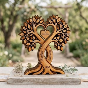 Op de afbeelding: Een houten boomsculptuur met twee verstrengelde stammen die een hartvorm vormen. De stammen zijn gegraveerd met de namen Sophia en Anthony en een oneindigheidssymbool. Een hartvormige hanger in het midden toont de datum 10.08.2026.