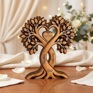 Op de afbeelding: Een gelaagd houten boomsculptuur met twee verstrengelde stammen waarin de namen Sophia en Anthony zijn gegraveerd, een hartvormige uitsparing in het midden en een hangend houten hartje met de datum 10.08.2026 en een oneindigheidssymbool.