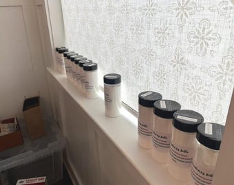 May include: A collection of white cylindrical containers with black lids, lined up on a windowsill. Each container has a label with text. The background features a patterned window covering and a glimpse of other items.