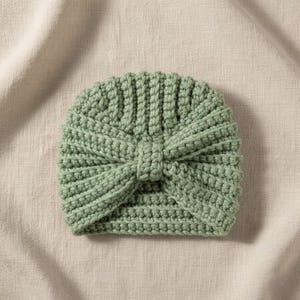 May include: A sage green crochet turban hat. The hat features a ribbed design on the top and a knotted bow detail at the front. The hat is made of soft yarn and is suitable for babies and children.