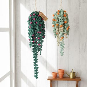 May include: Two hanging crochet plant decorations. One is dark green with pink accents, the other is light green with orange accents. Both are suspended by twine. A small wooden shelf holds terracotta pots and a brass watering can.